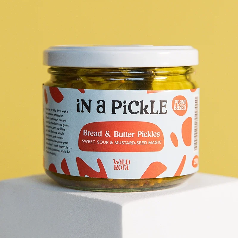 A glass jar of Wild Root 'In a PickLE' Bread & Butter Pickles sits on a white display pedestal against a solid yellow background.