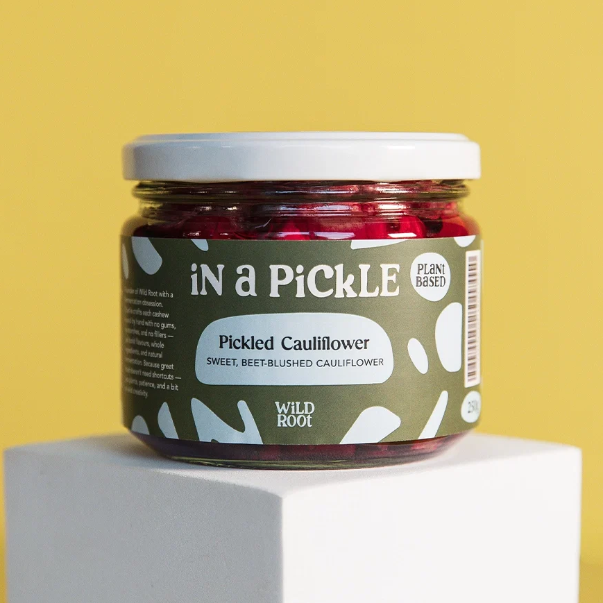 A jar of Wild Root 'In a Pickle' Pickled Cauliflower, featuring a dark olive green label with white accents and a white lid, displayed on a white pedestal against a yellow background.