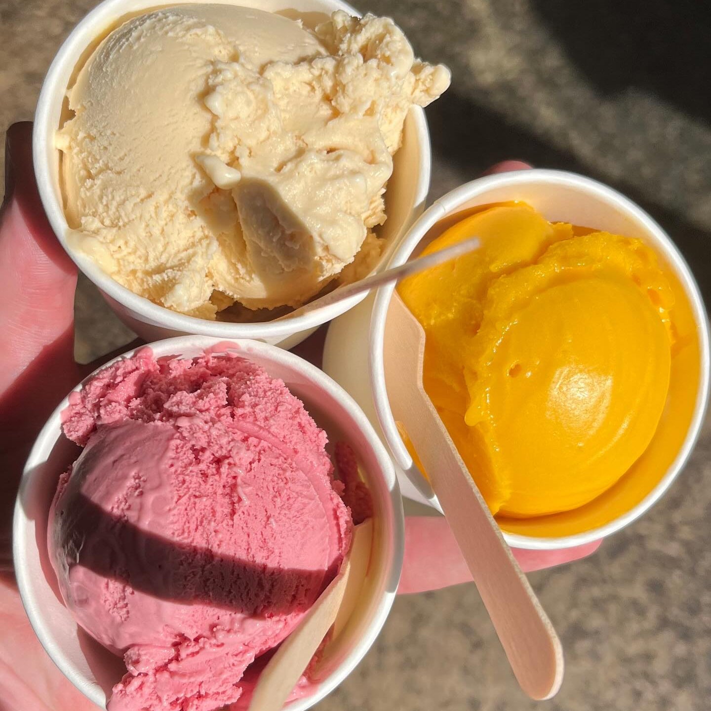Three small paper cups of ice cream held in a hand outdoors in bright sunlight: vanilla ice cream, pink strawberry ice cream, and yellow mango ice cream, each with a wooden spoon.