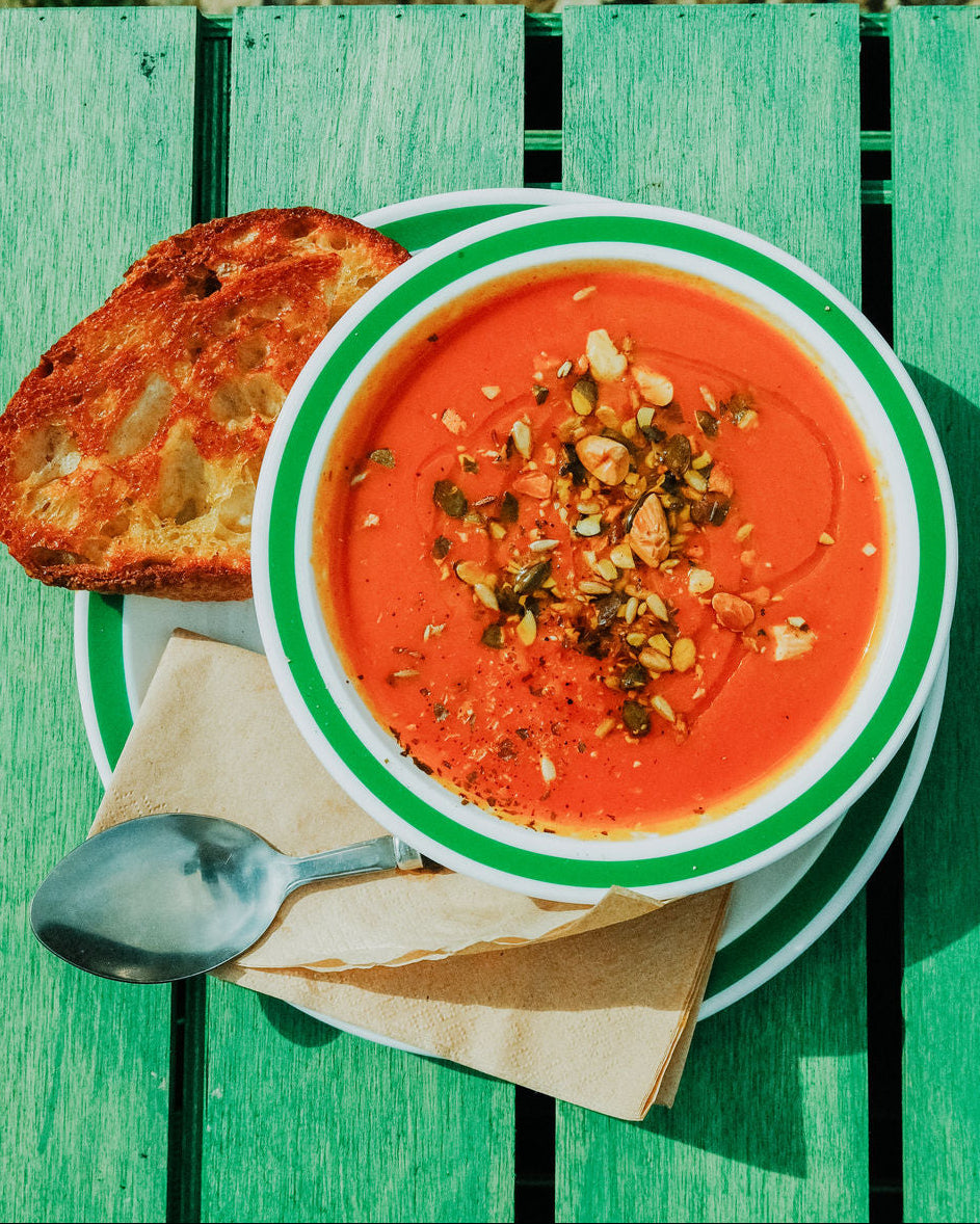 A bowl of vibrant orange-red soup with green-rimmed edges, topped with nuts and spices, served on a green wooden table alongside a slice of toasted bread, a spoon, and a napkin.