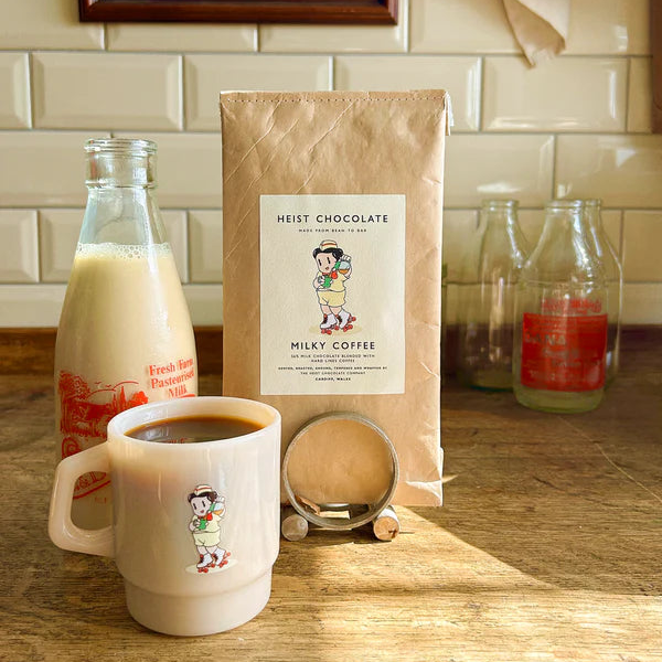 Coffee mug, milk bottle, and Heist Milky Coffee chocolate package on a wooden surface with a tiled wall background