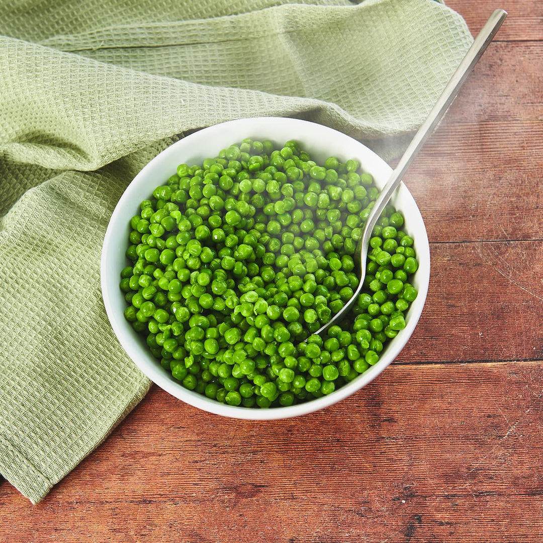 White bowl filled with green peas on a wooden surface with a green cloth.