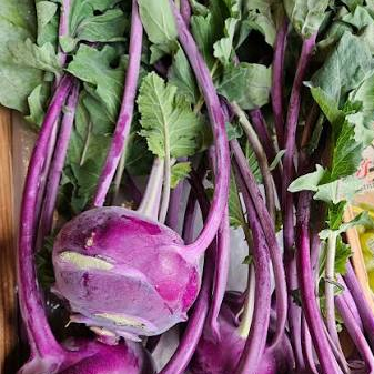 Several purple kohlrabi vegetables with long green leaves and stems, arranged on a wooden tray with other produce partially visible around them.